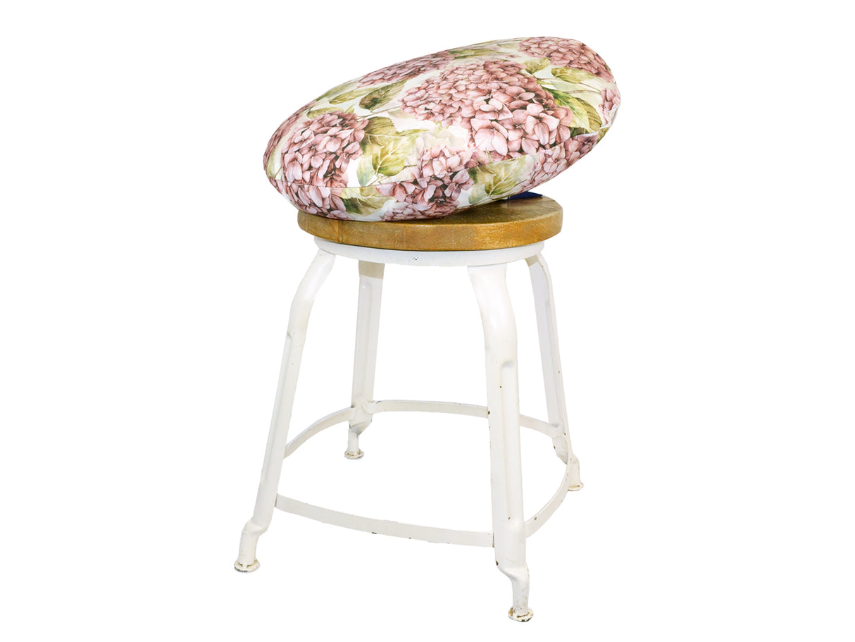 Auf einem Hocker liegt ein runder Kissenbezug im Landhausstil, bedruckt mit rosanen/pinken Hortensien-Blueten.