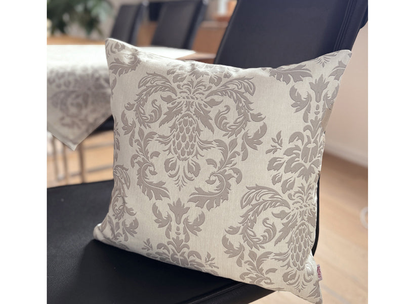 Auf einem schwarzen Lederstuhl, lehnt ein Kissen, bezogen mit einem beigen Kissenbezug der mit barocken Ornamenten in einem dunkleren Beige, bedruckt ist.