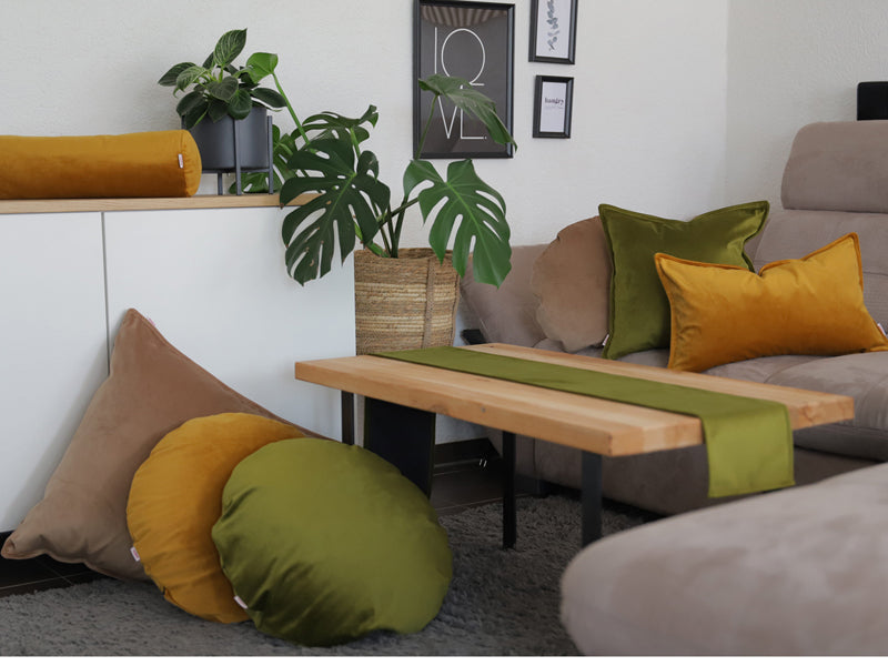 Curry-senf-gelbe, braun-beige und mai-gruene Samt Kissenbezuege und ein mai-gruenes Tischband sind in einem Wohnzimmer dekoriert.