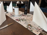 Weisser Tischlaeufer mit gefrosteten Rosen in rosa, Kieferzapfen und Zweigen aus der beties Magie des Winters Kollektion "Frost-Rose" auf einem gedeckten Holztisch.