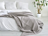 In einem Schlafzimmer liegt eine Nebel-graue Tagesdecke aus der beties "PUR.NATURE" Serie auf einem Bett ausgebreitet.